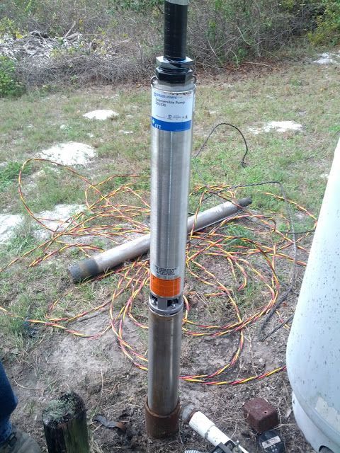 A stainless steel submersible well pump assembly standing vertically on the ground outdoors next to electrical wiring.