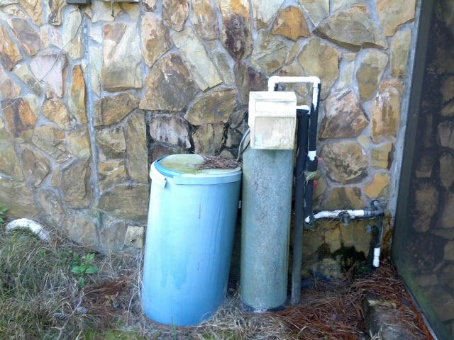 A blue brine tank next to a tall, grey water softener tank with white and black piping against a stone wall.