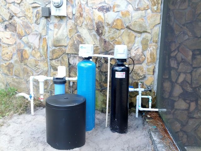 A water filtration system featuring a blue tank, black tank, and plumbing installed against a stone wall outdoors.