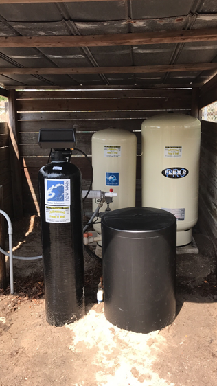 Water treatment system with a black filter, brine tank, and two beige pressure tanks inside an outdoor wooden shed.