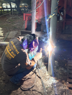 A technician wearing a welding helmet and protective gloves works on a metal pole, creating a bright flare of light.