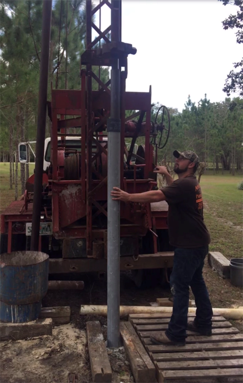 A person guides a steel pipe into the ground while operating a mobile drilling rig in an outdoor, wooded area.