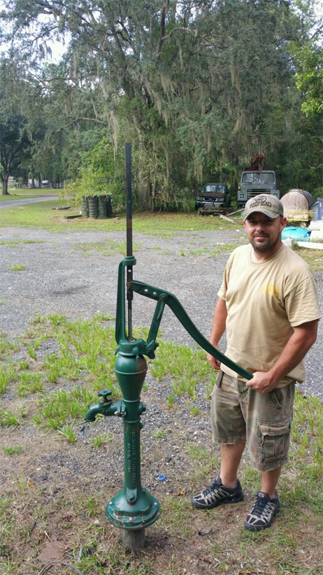 A person stands outdoors next to a tall, green, antique manual water pump installed in a gravel lot.
