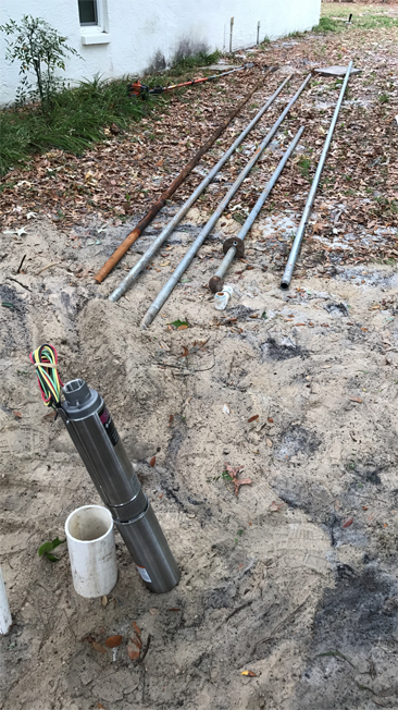 A metal submersible water well pump and several sections of pipe resting on sandy ground outside a building.