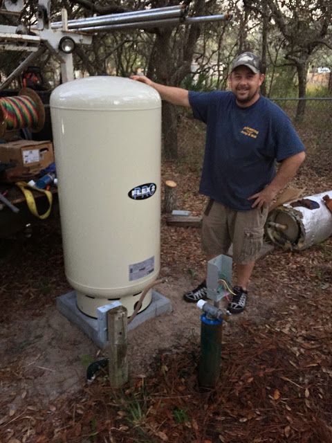 A smiling technician stands next to a large cream-colored well water pressure tank installed outdoors on a concrete base.
