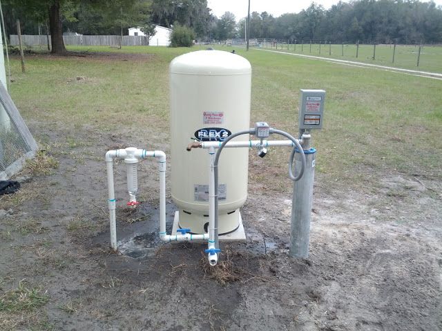 A beige well pressure tank and associated PVC plumbing and electrical disconnect box located outdoors on grass.
