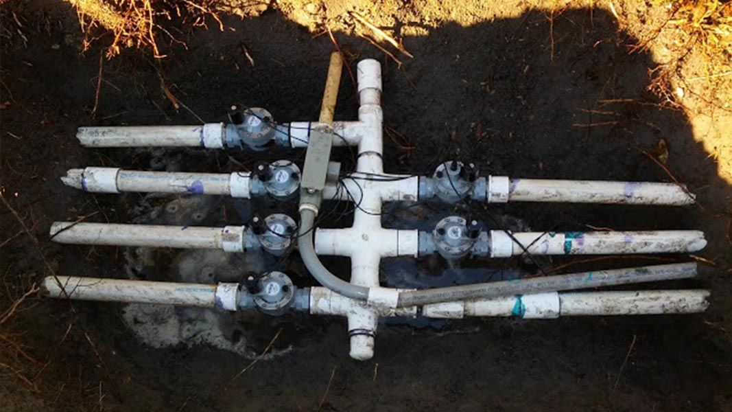A multi-zone irrigation valve manifold with white PVC pipes connected to electric solenoid valves in a dirt pit.