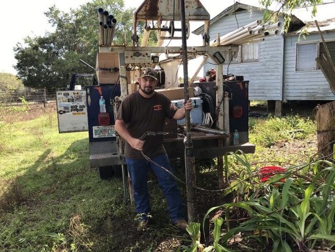 A man in a brown t-shirt and jeans stands next to a well-drilling rig parked in a grassy yard beside a small house.
