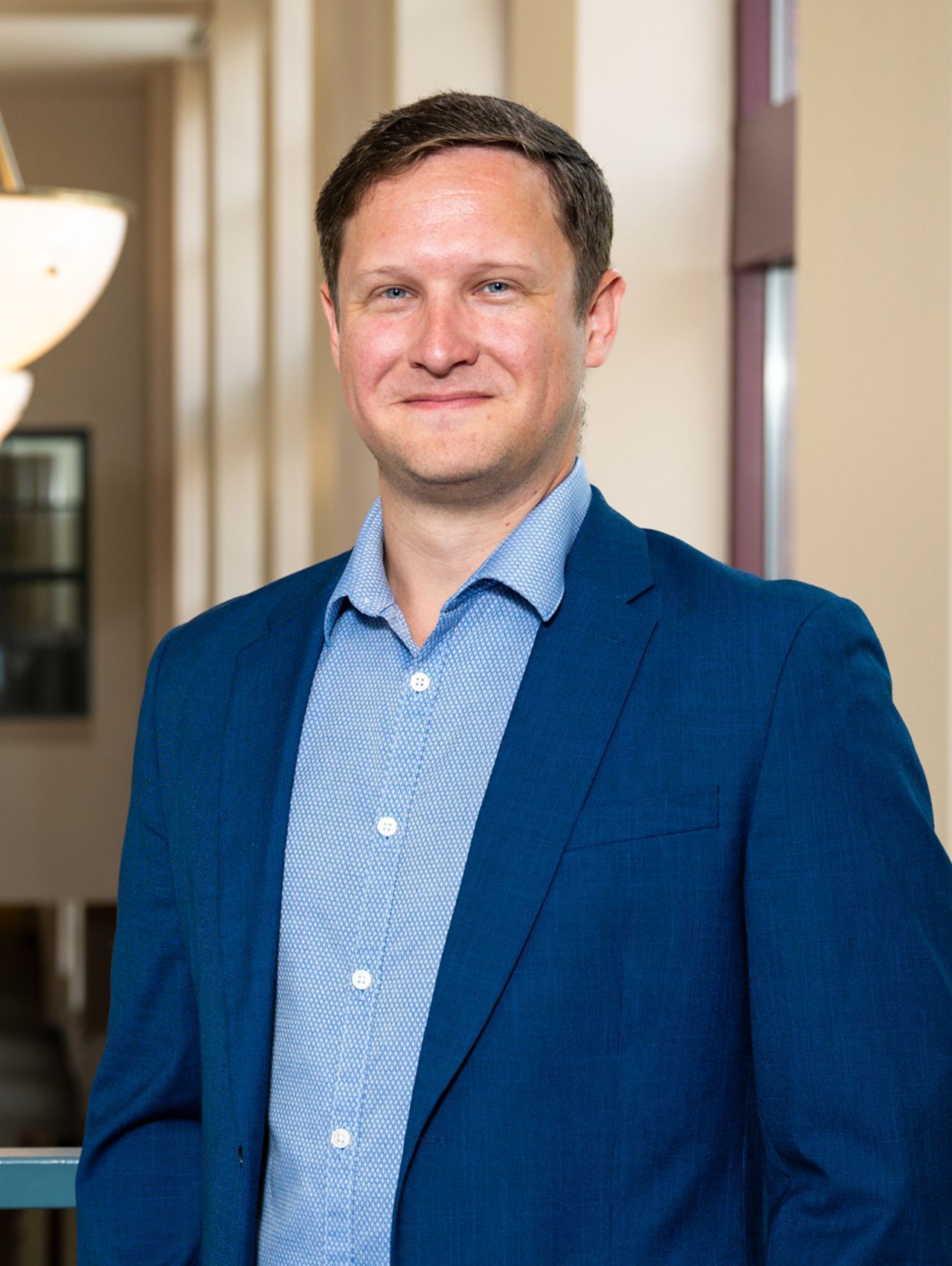 Man in a blue blazer standing indoors, smiling slightly