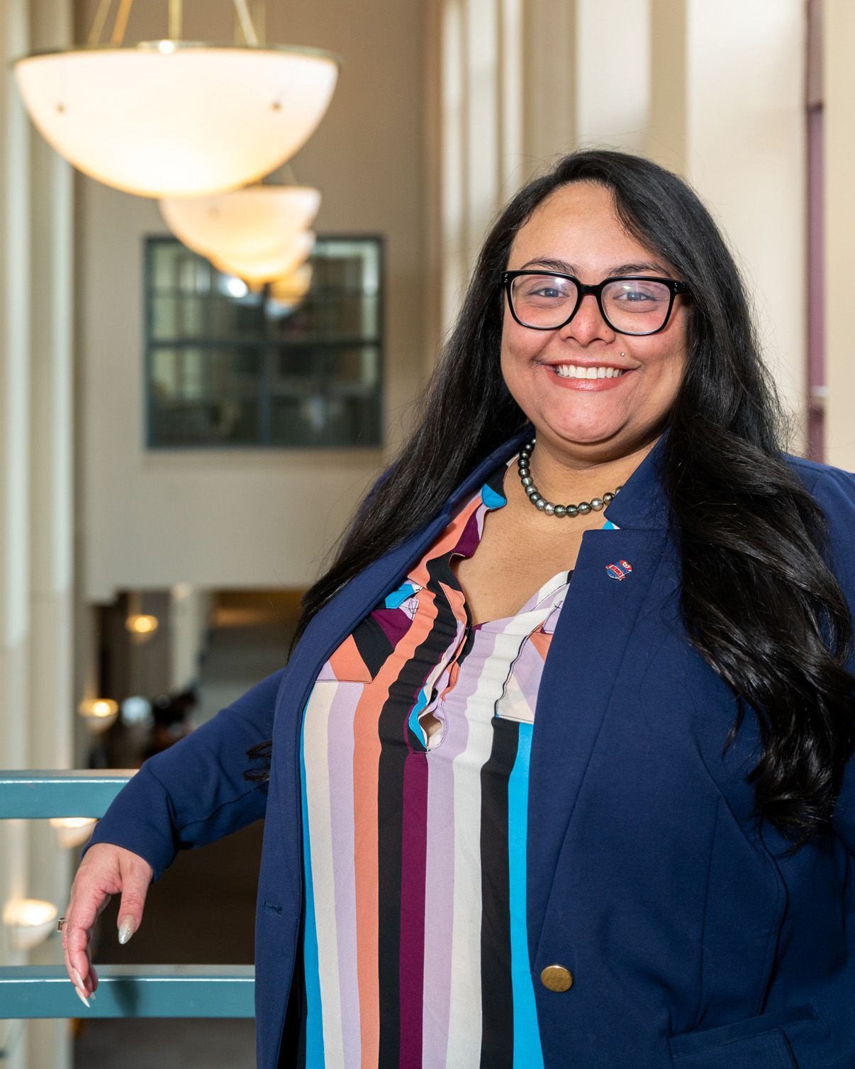 Smiling person in glasses and blue blazer standing indoors beside a railing and hanging lights