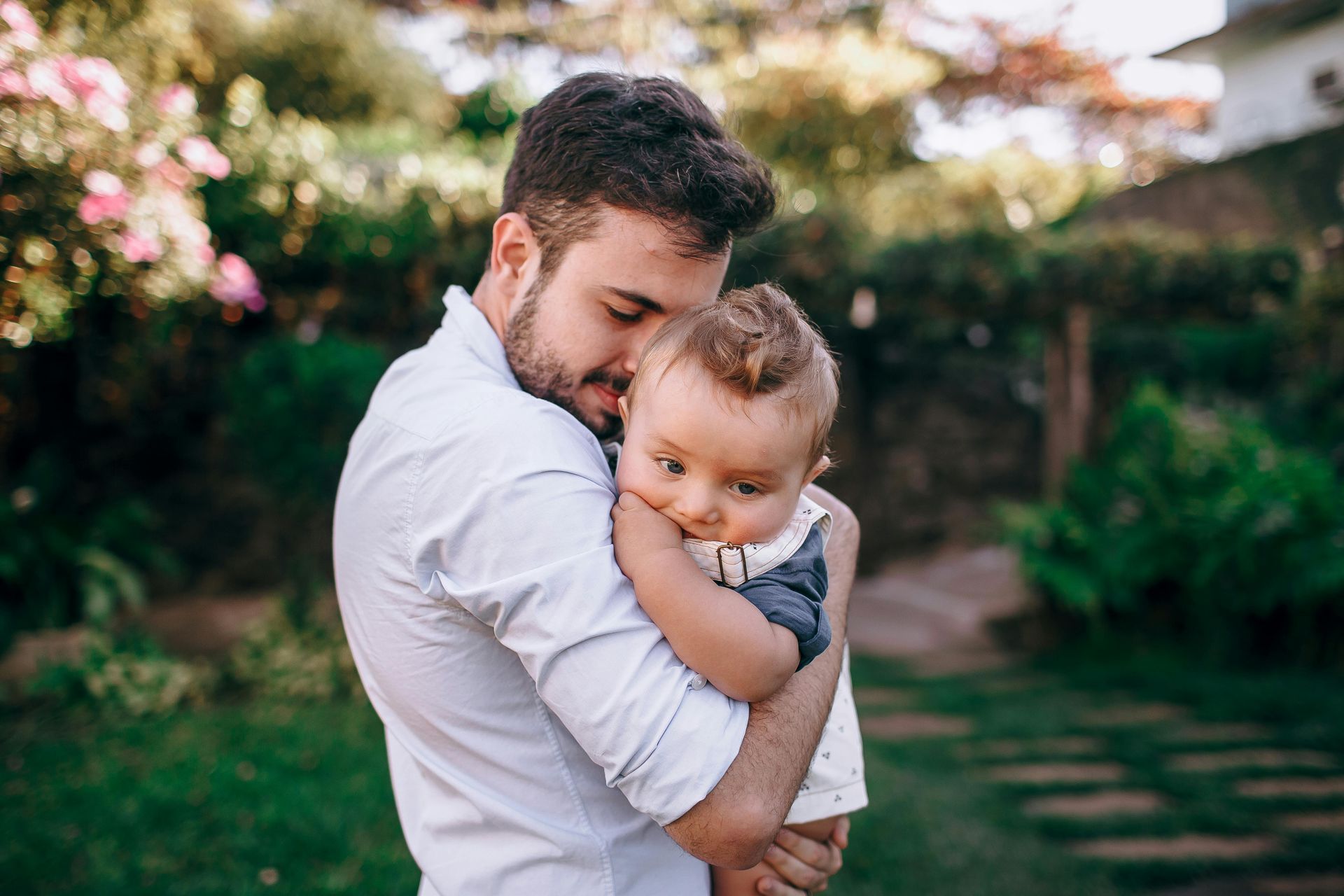 Man in a white shirt holding a baby outdoors in a garden, both looking calm.