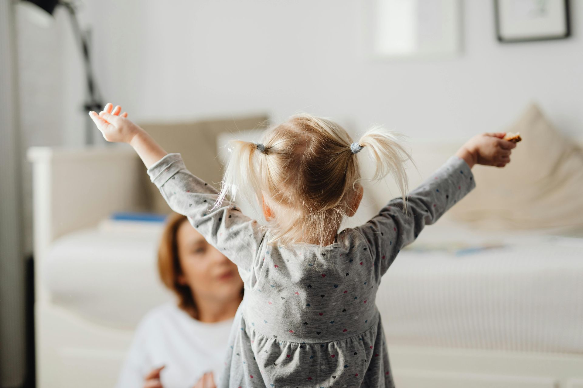 Child with arms raised in a bright bedroom, facing a smiling adult on a bed.