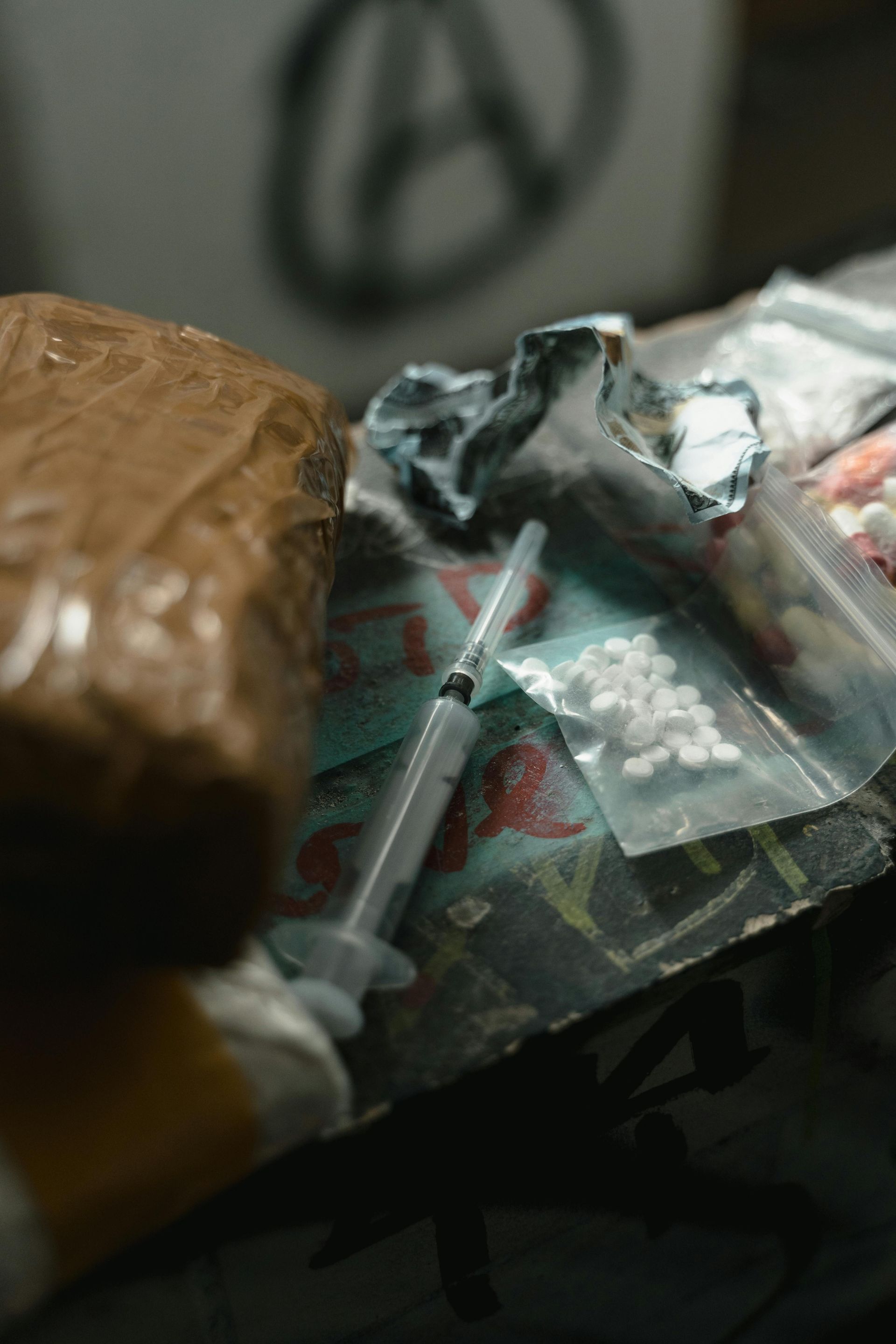 A cluttered tabletop with a syringe, plastic bags, and assorted pills or drug paraphernalia.