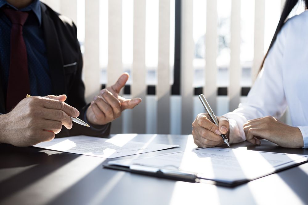 Two people in business attire discussing paperwork at a bright office table, one writing notes.