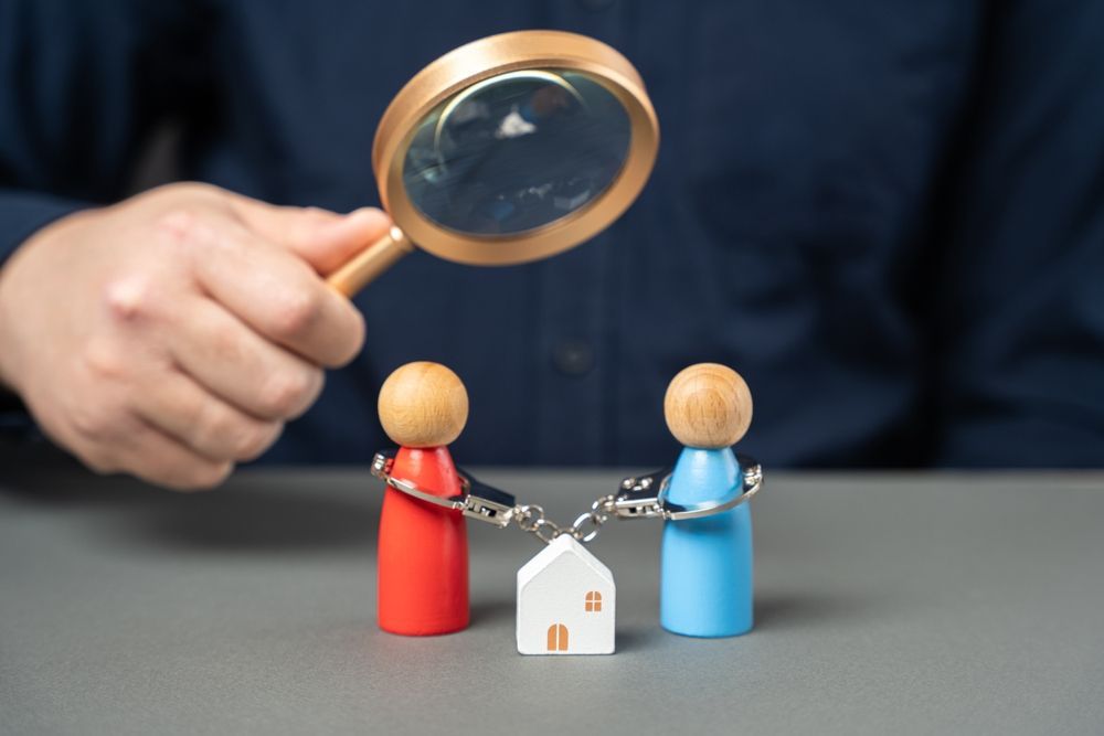 Magnifying glass over two wooden figures and a small house connected by a chain on a table