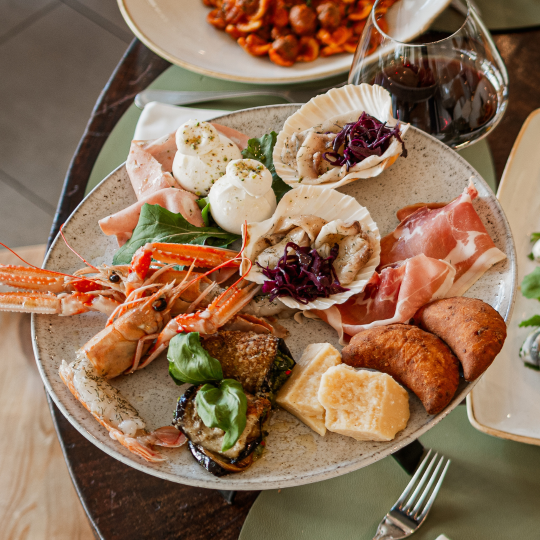 Assiette d'amuse-gueules avec fruits de mer, prosciutto, fromage et pâtes, accompagnée d'un verre de vin rouge.