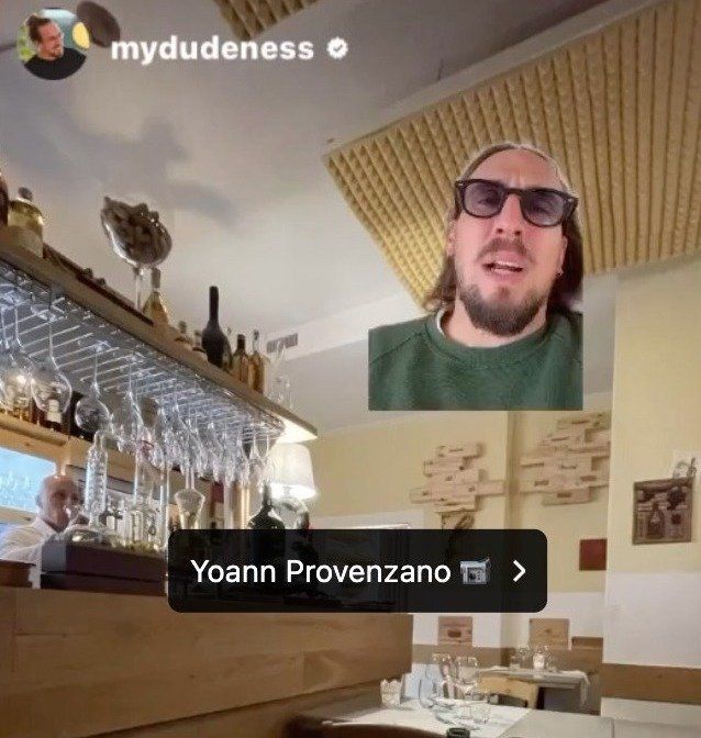 Un homme portant des lunettes de soleil parle dans un restaurant où l'on aperçoit le bar et un serveur, photographie un utilisateur d'Instagram.