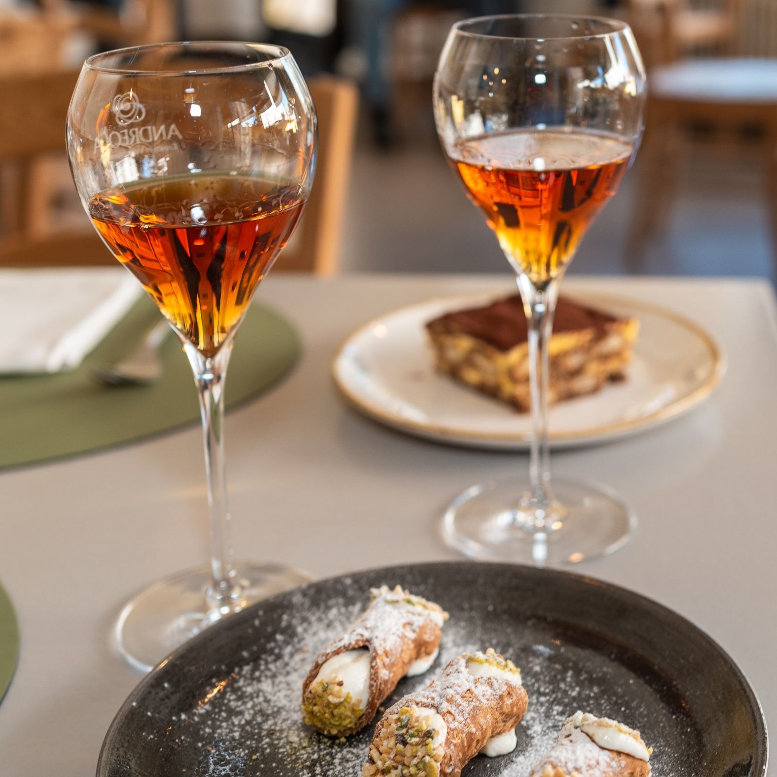 Deux verres de liquide ambré, du tiramisu et des cannoli sur une table.