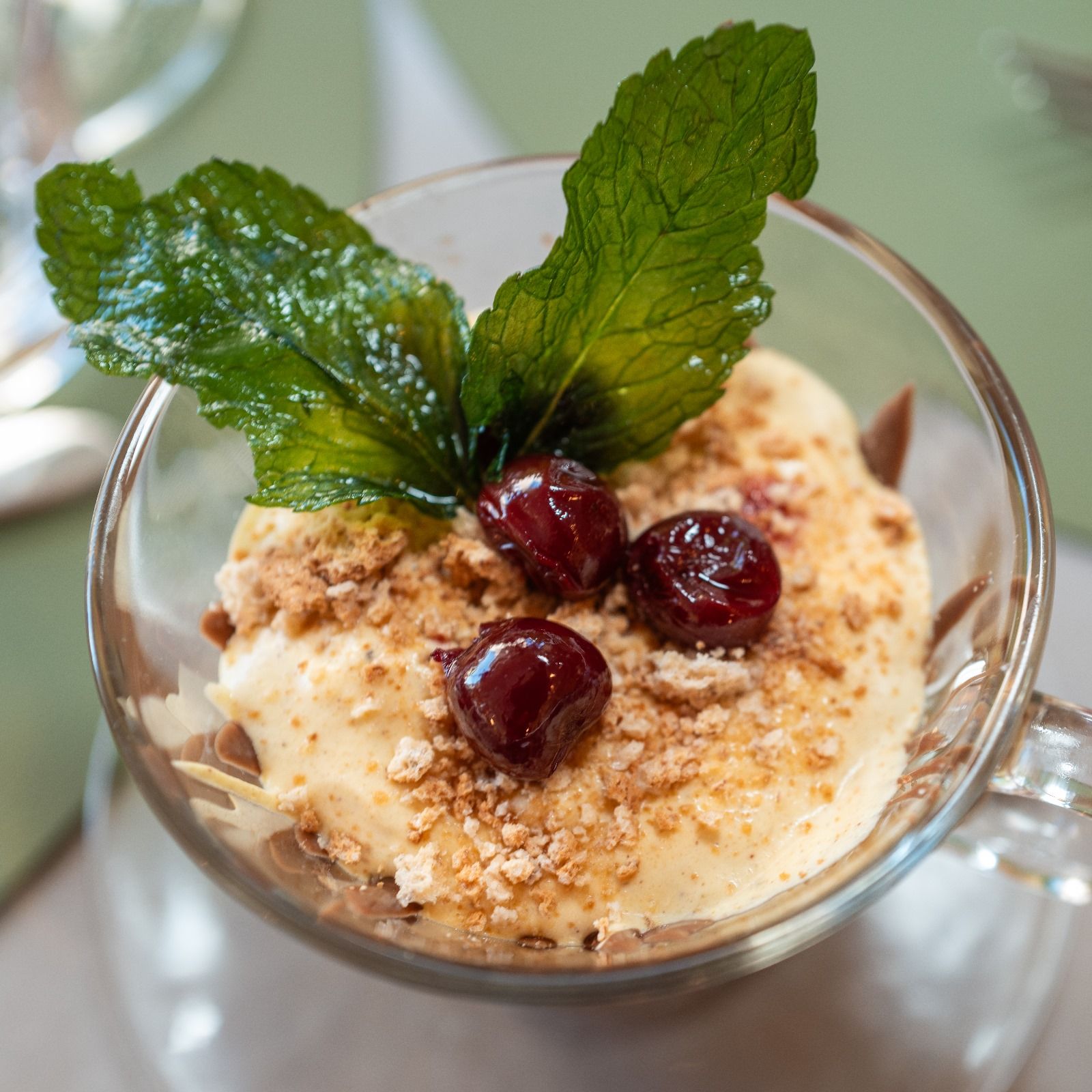 Dessert en verre : crémeux, garni de cerises, de miettes et d'un brin de menthe.