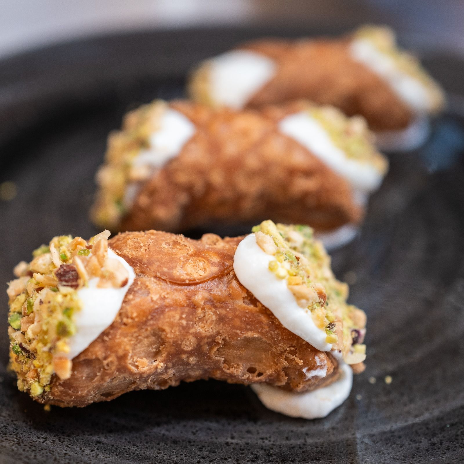 Trois cannoli fourrés à la crème blanche et aux noix hachées, présentés sur une assiette sombre.