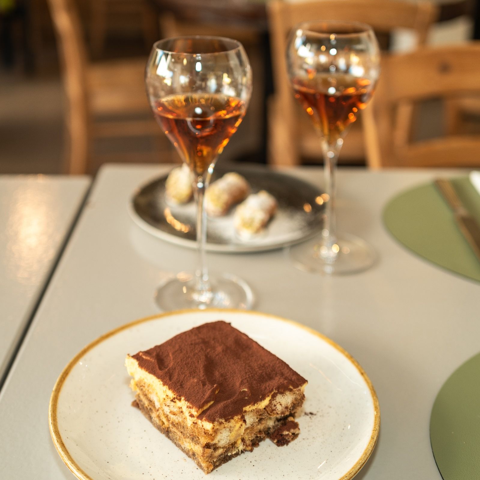 Tiramisu accompagné de deux verres de liqueur ambrée sur une table ; petites pâtisseries en arrière-plan.