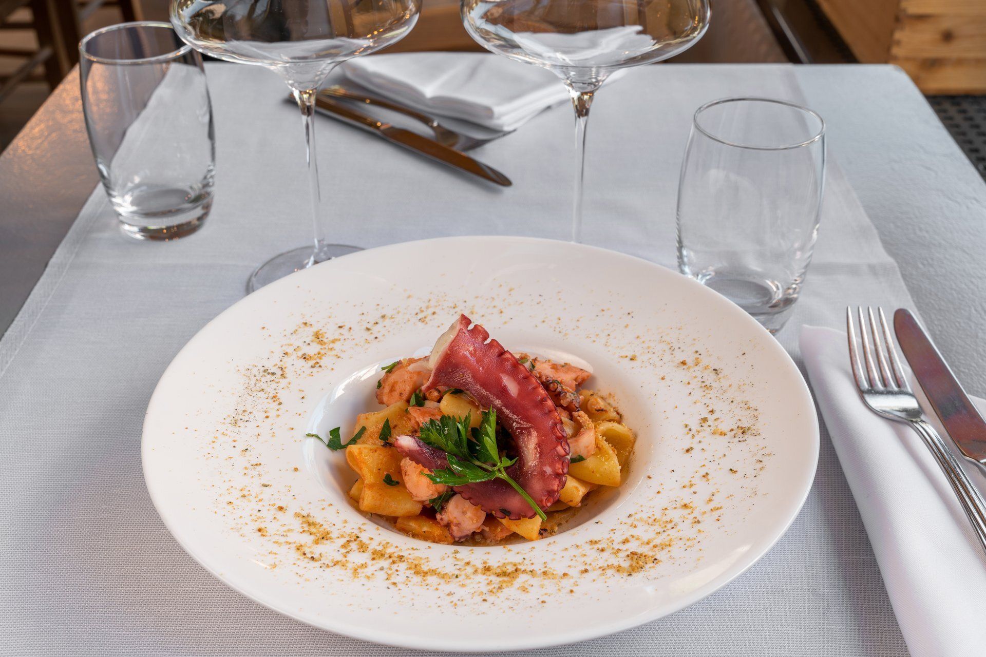 Plat de pâtes sur une assiette blanche, garni de poulpe et de persil, sur une table dressée avec des verres à vin et des couverts.