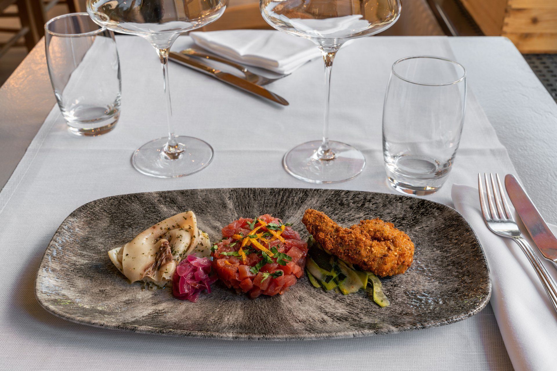 Assiette d'amuse-bouche avec trois échantillons, verres à vin et couverts sur une nappe blanche.