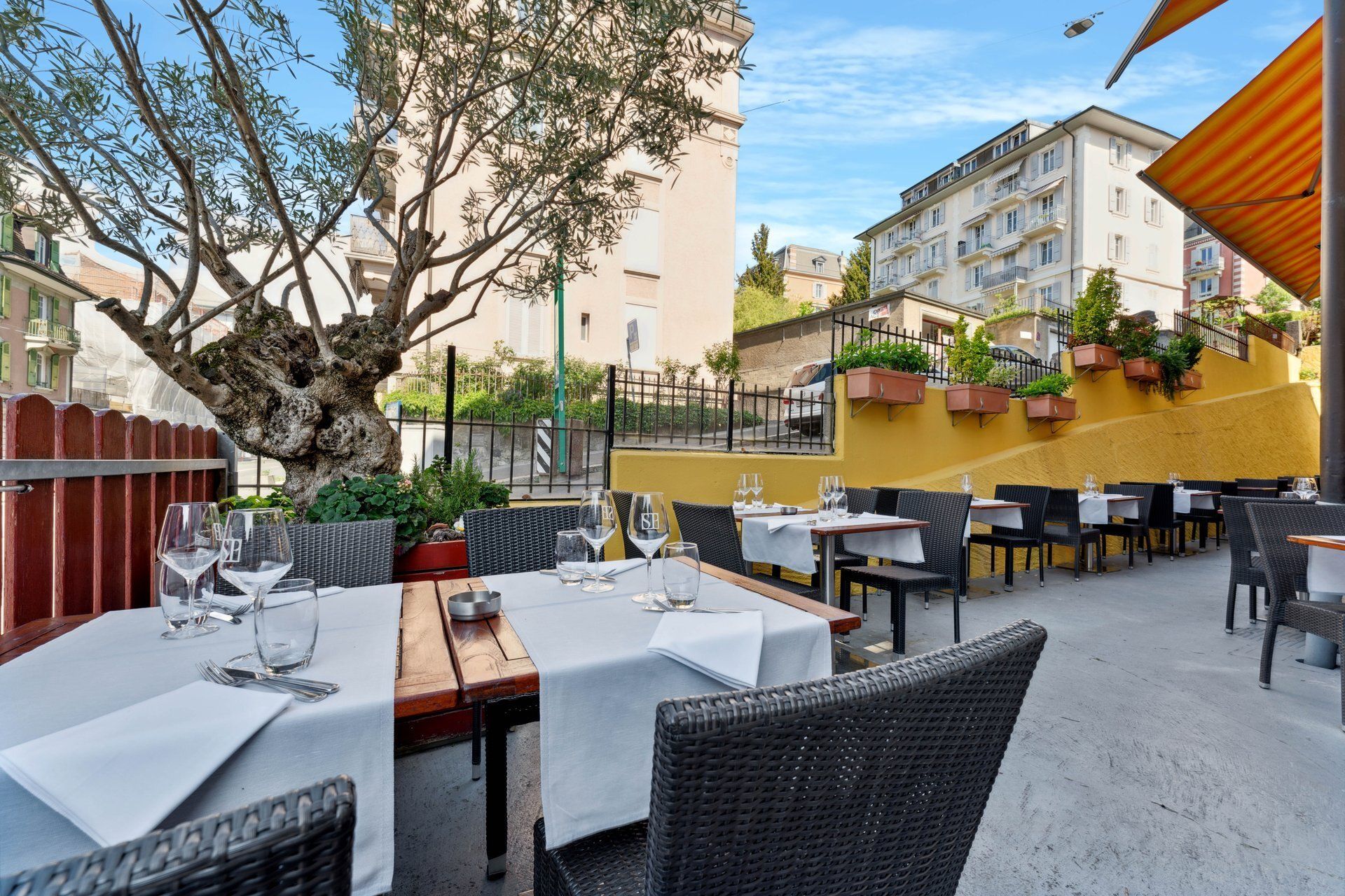 Restaurant en plein air avec tables dressées, mur jaune et bâtiment en arrière-plan.