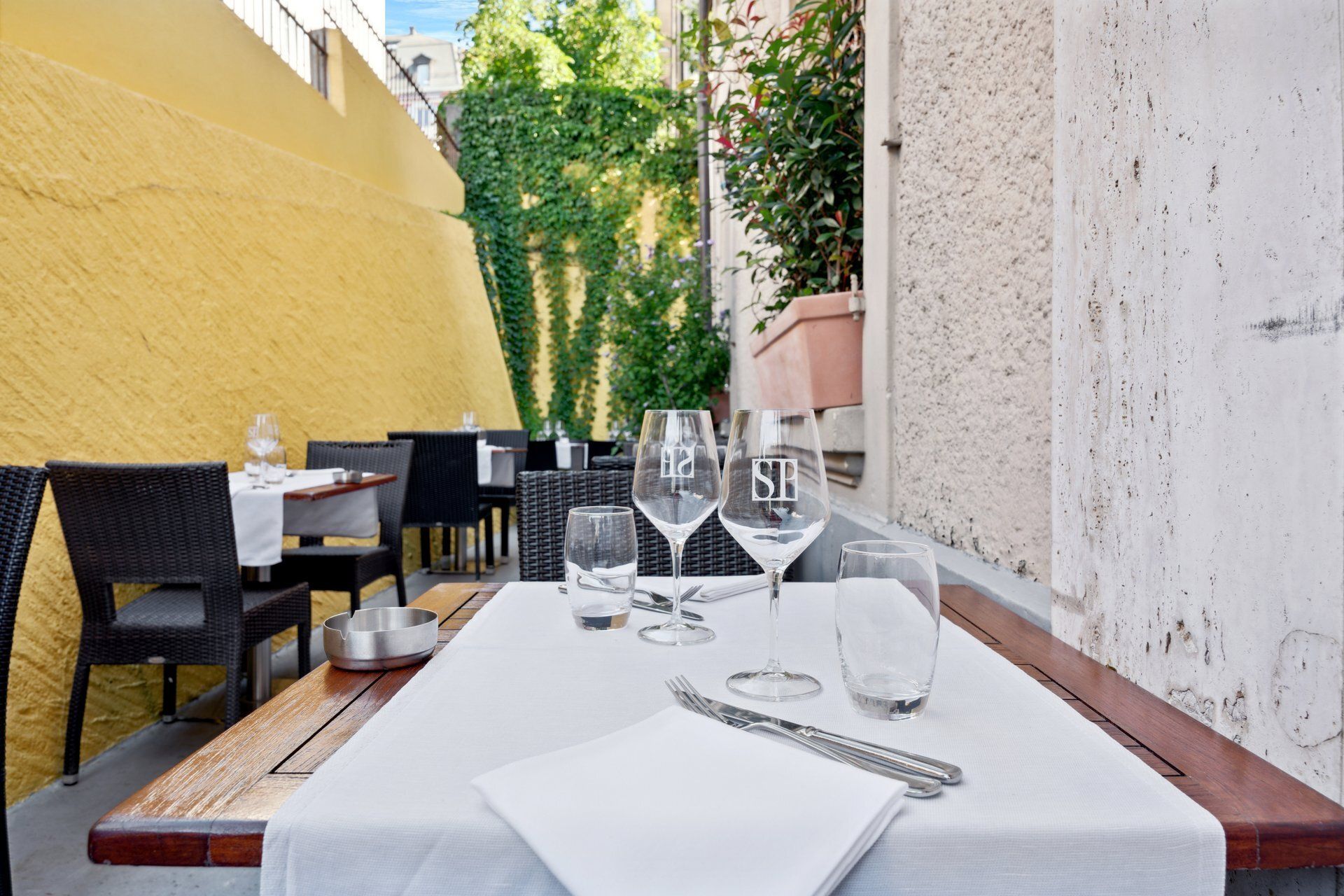 Table dressée en terrasse avec verres à vin et couverts. Mur jaune et verdure en arrière-plan.