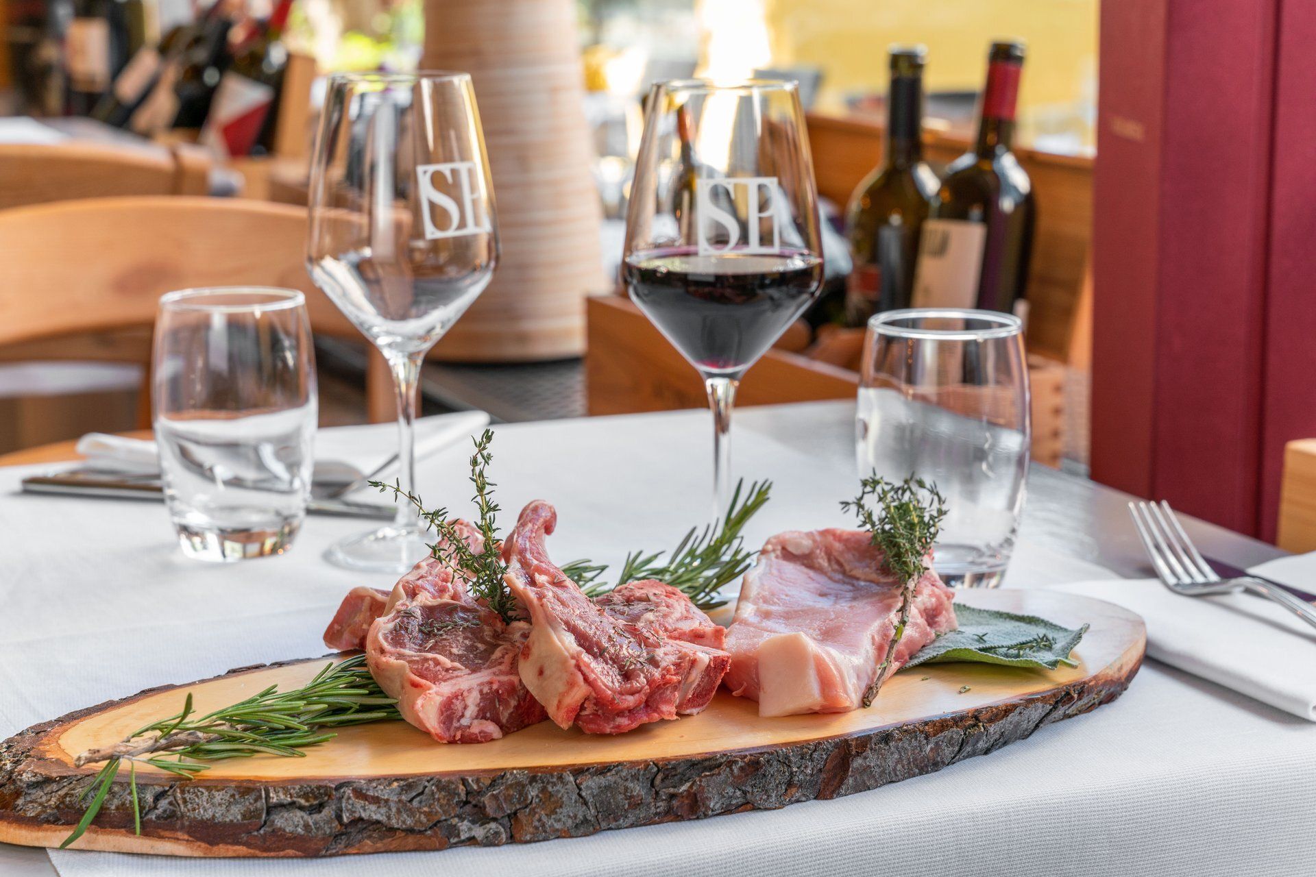 Carré d'agneau sur un plat en bois, avec des verres à vin et des bouteilles sur une table.