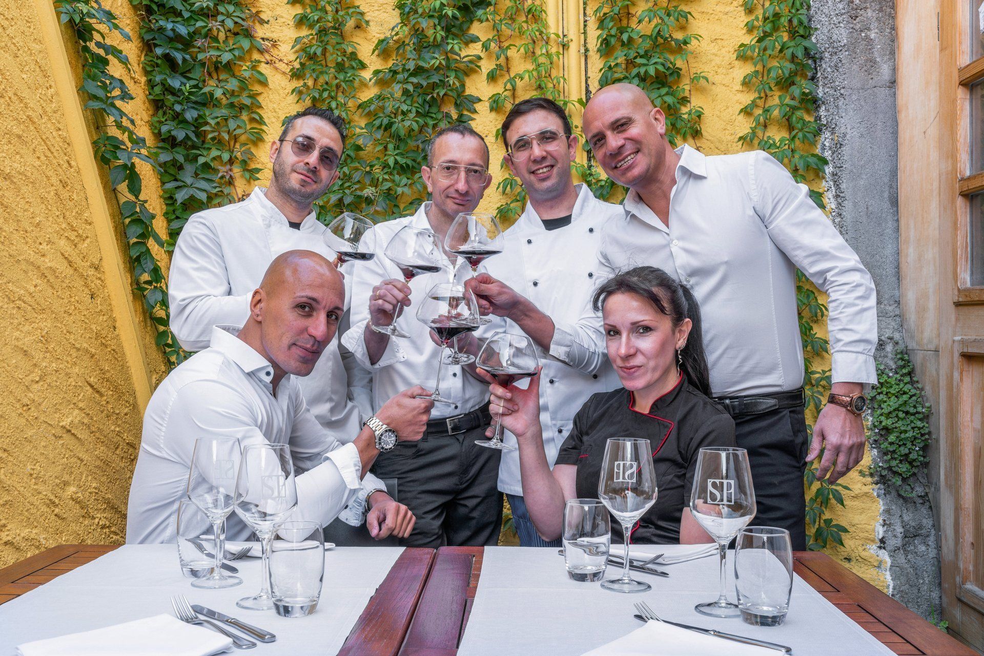 Des chefs trinquent avec leurs verres de vin à une table, souriants, devant un mur jaune orné de verdure.