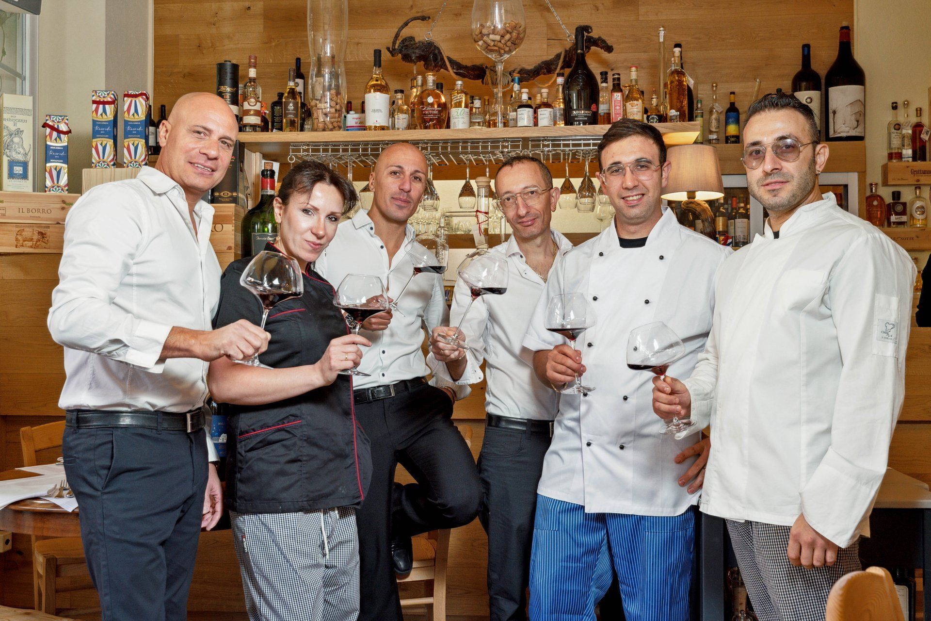 Six employés de restaurant, verres à vin à la main, sourient devant un bar où sont disposées des bouteilles.
