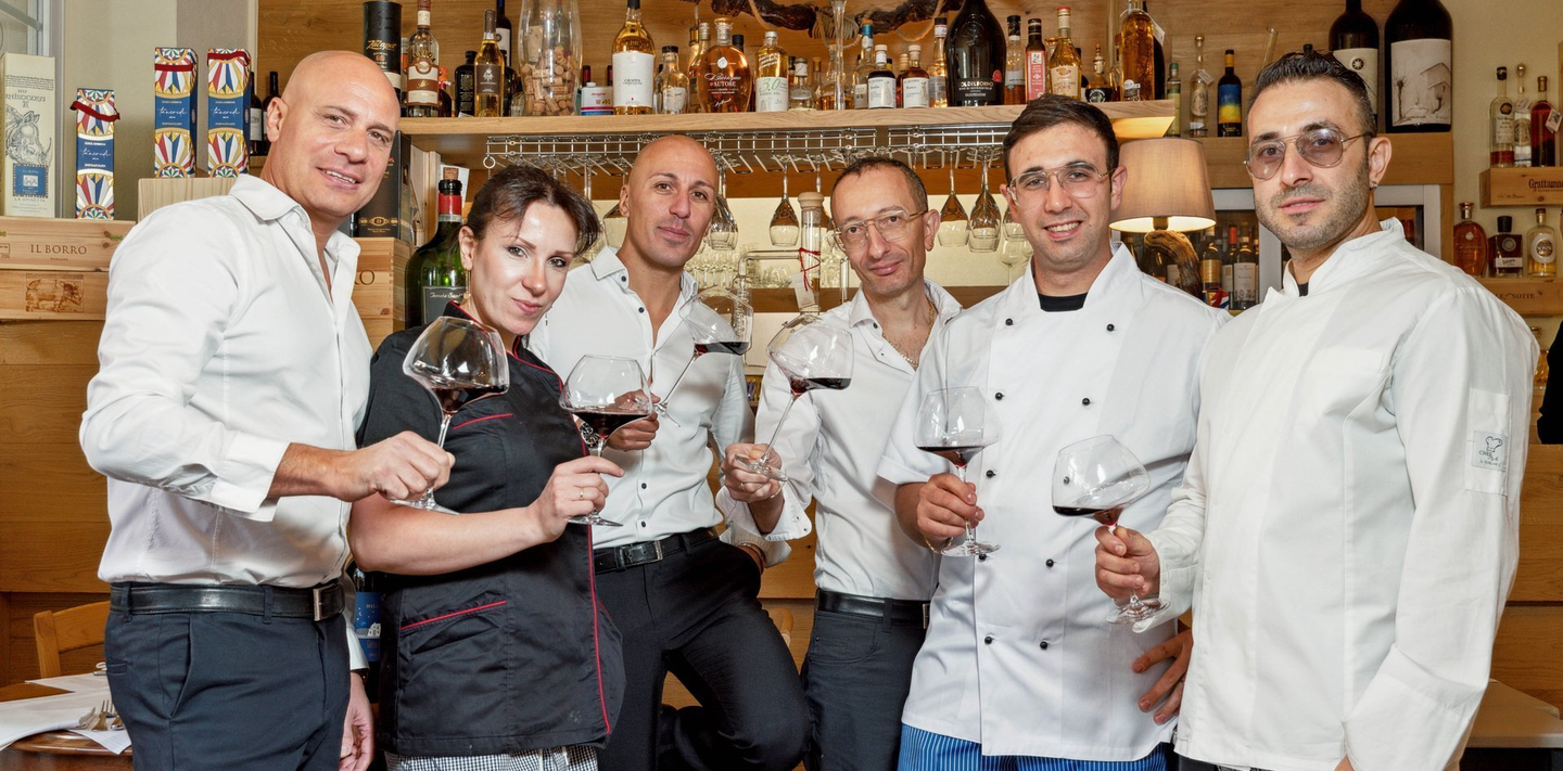 Six employés de restaurant, verres de vin rouge à la main, sourient.