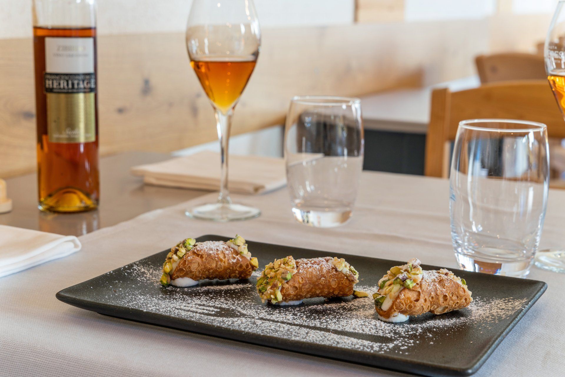 Trois cannoli sur une assiette noire, saupoudrés de sucre glace, près de verres à vin et d'une bouteille sur une table.