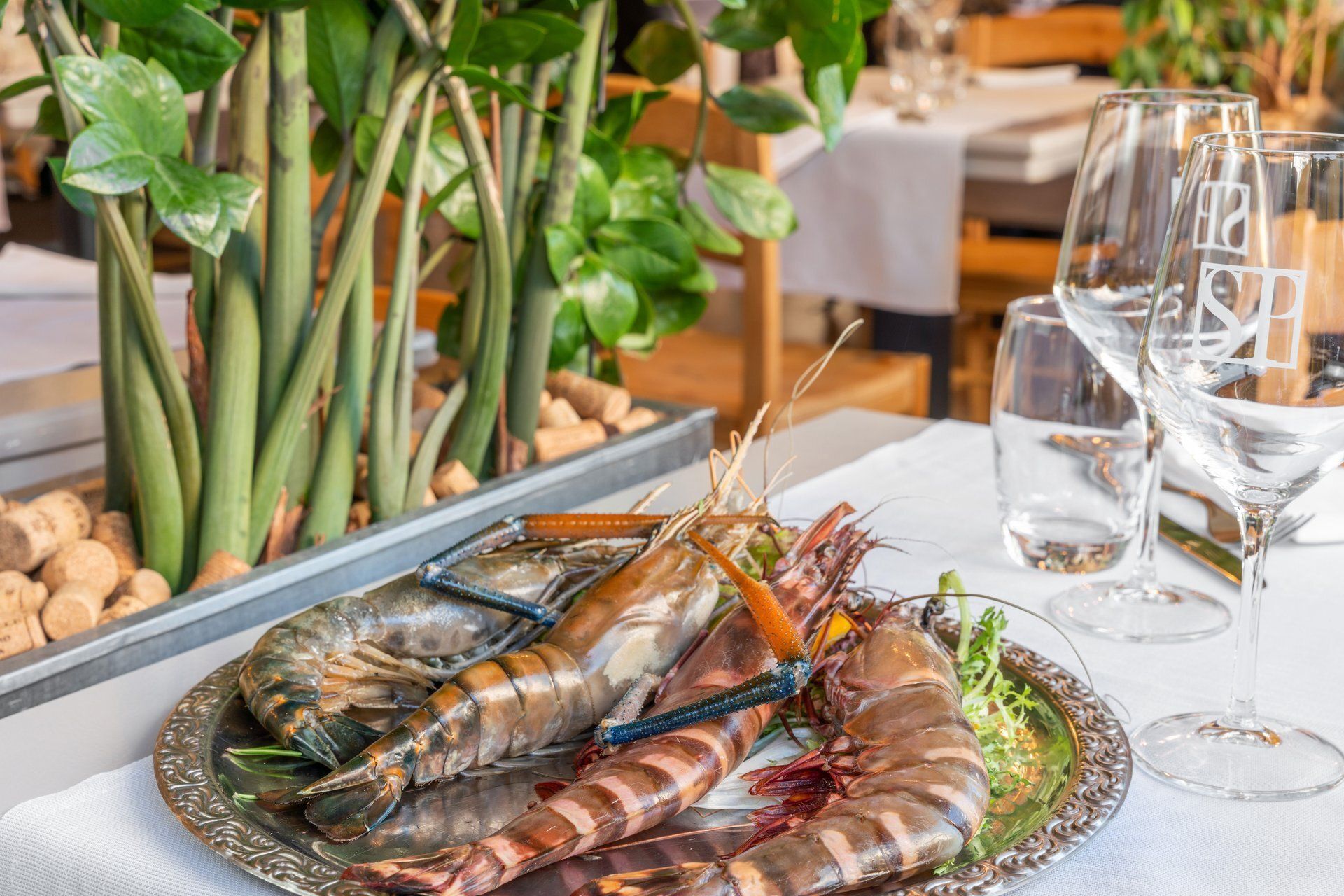 Des crevettes grillées sur un plat en argent, posées sur une nappe blanche dans un restaurant décoré de plantes et de verres.