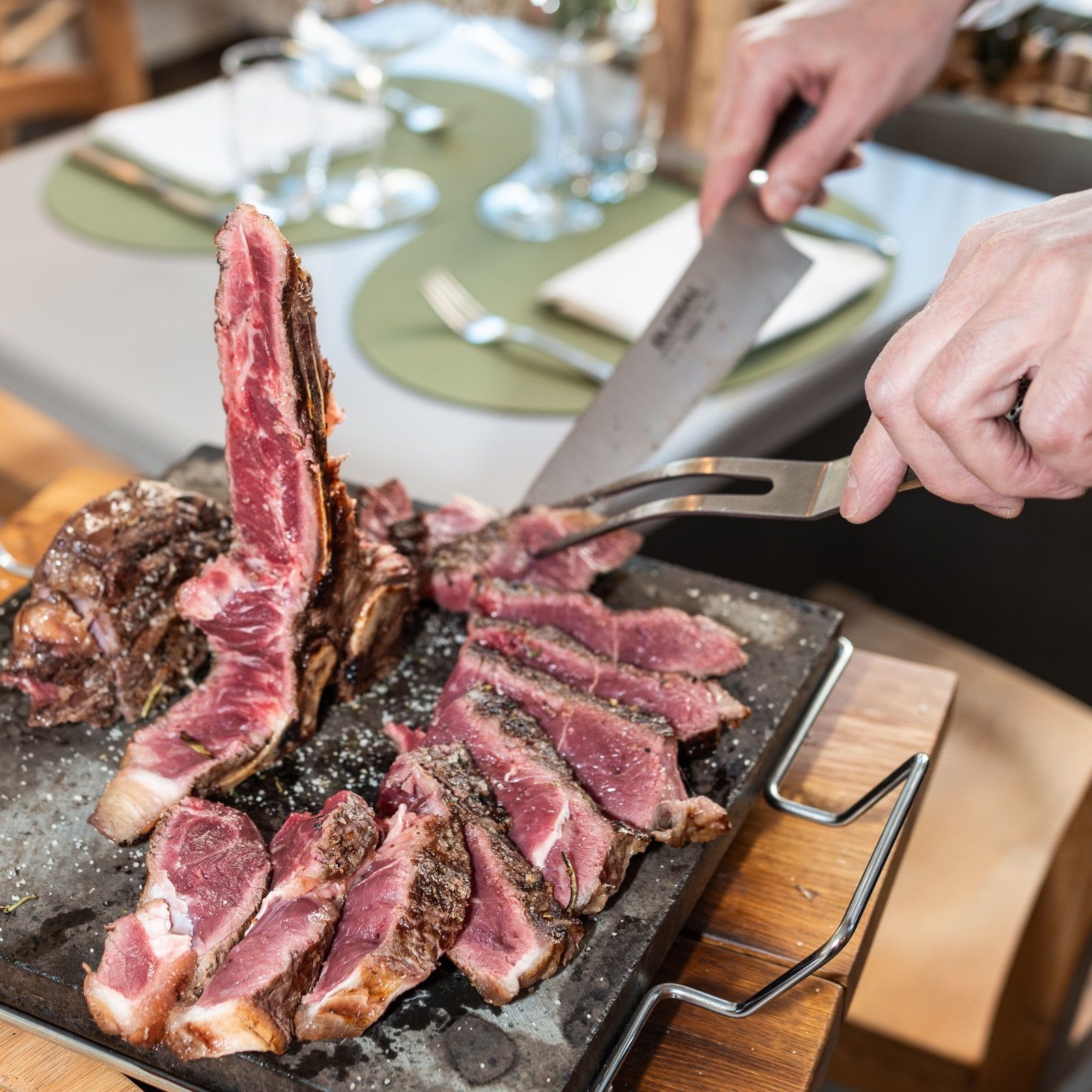 Une personne découpe un steak cuit sur une pierre chaude dans un restaurant. Une table est dressée en arrière-plan.
