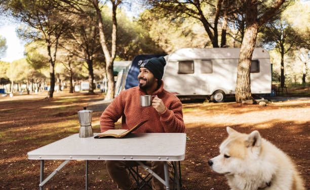 Man drinking coffee at campsite table with dog and RV.
