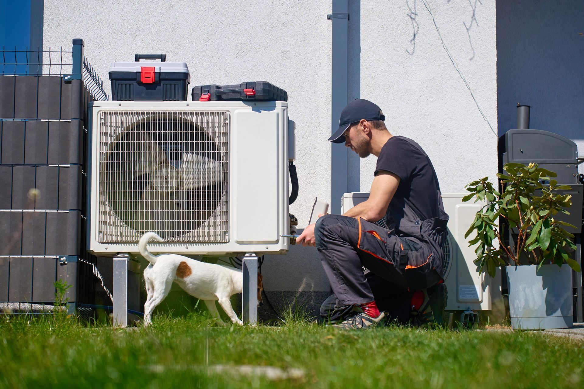 Technician kneels beside a white outdoor HVAC unit while a small white dog stands nearby in a yard. Technician kneels beside a white outdoor HVAC unit while a small white dog stands nearby in a yard.