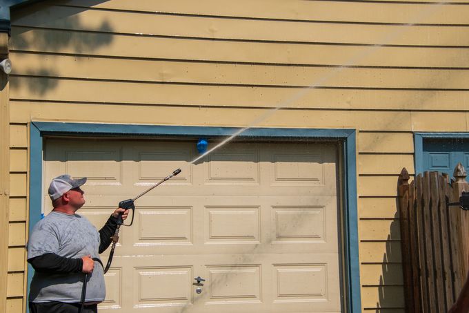 Person power-washing a garage door and siding on a house with a long spray wand