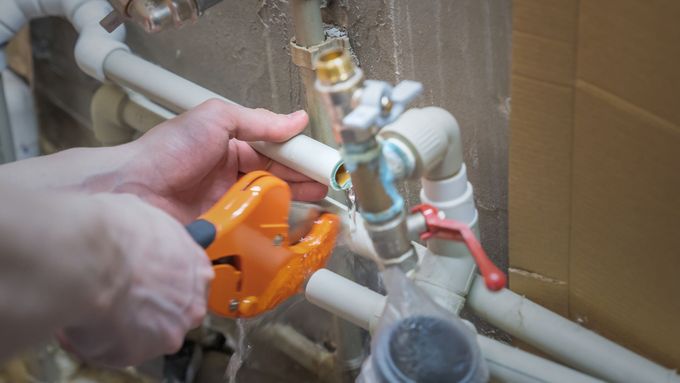 Hands tightening orange pipe cutter on white plumbing pipes under a sink.