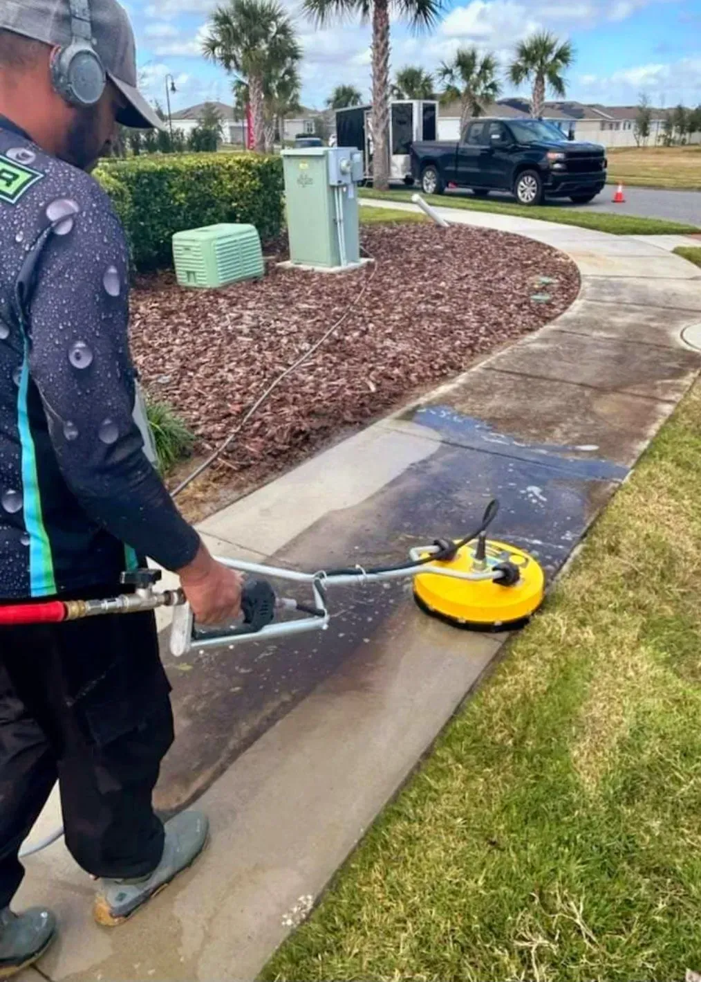 Worker cleaning a sidewalk with a yellow surface cleaner near a landscaped curb on a sunny day