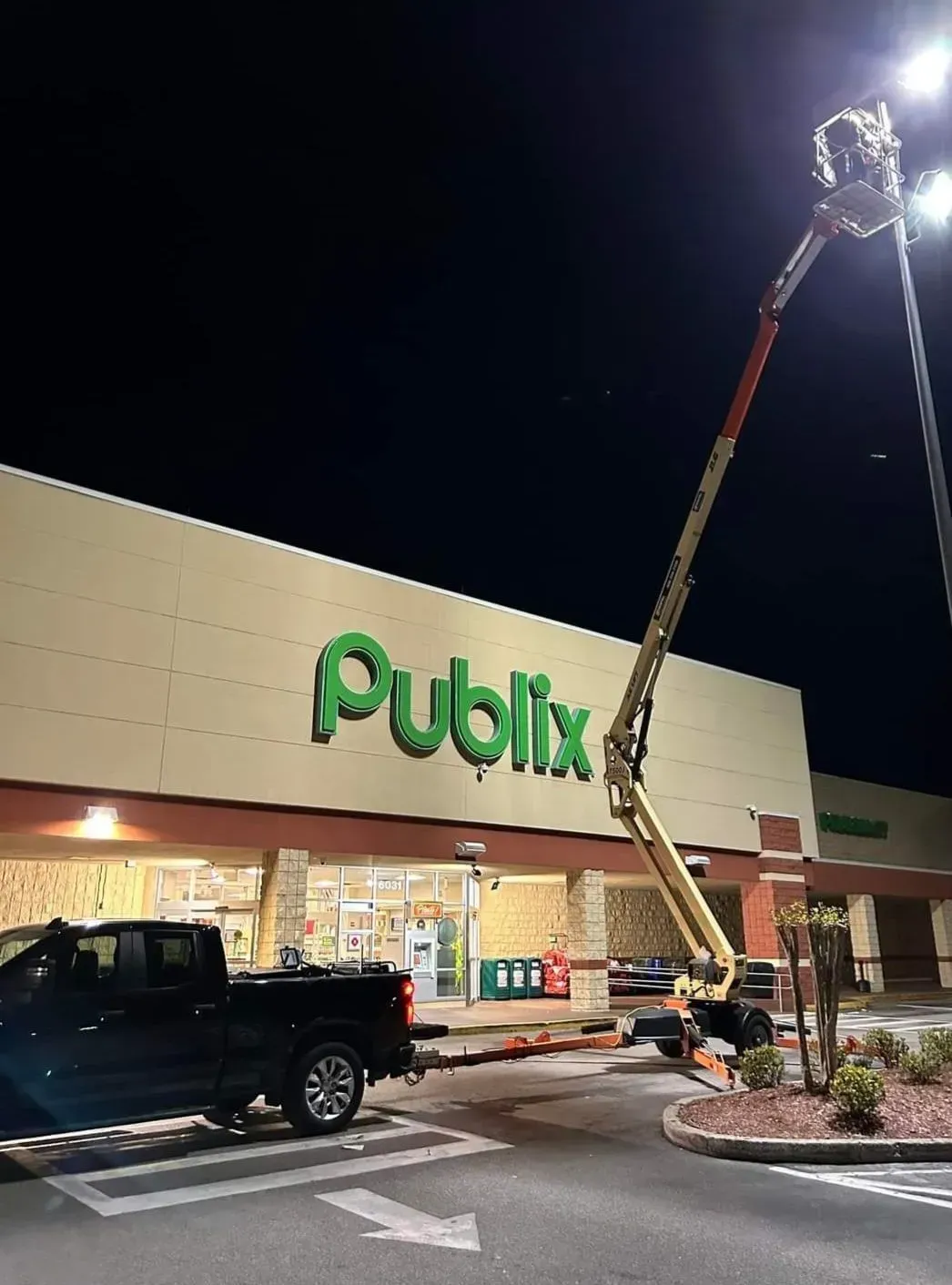 Publix storefront at night with a raised bucket truck and worker repairing the roof sign