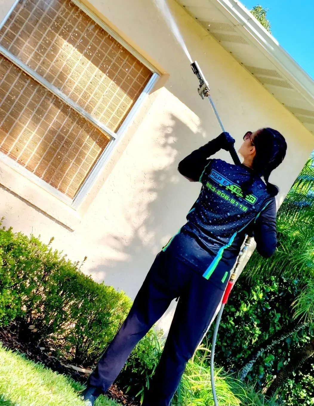 Person using a long-handled tool to clean the outside of a house beside a window and bushes