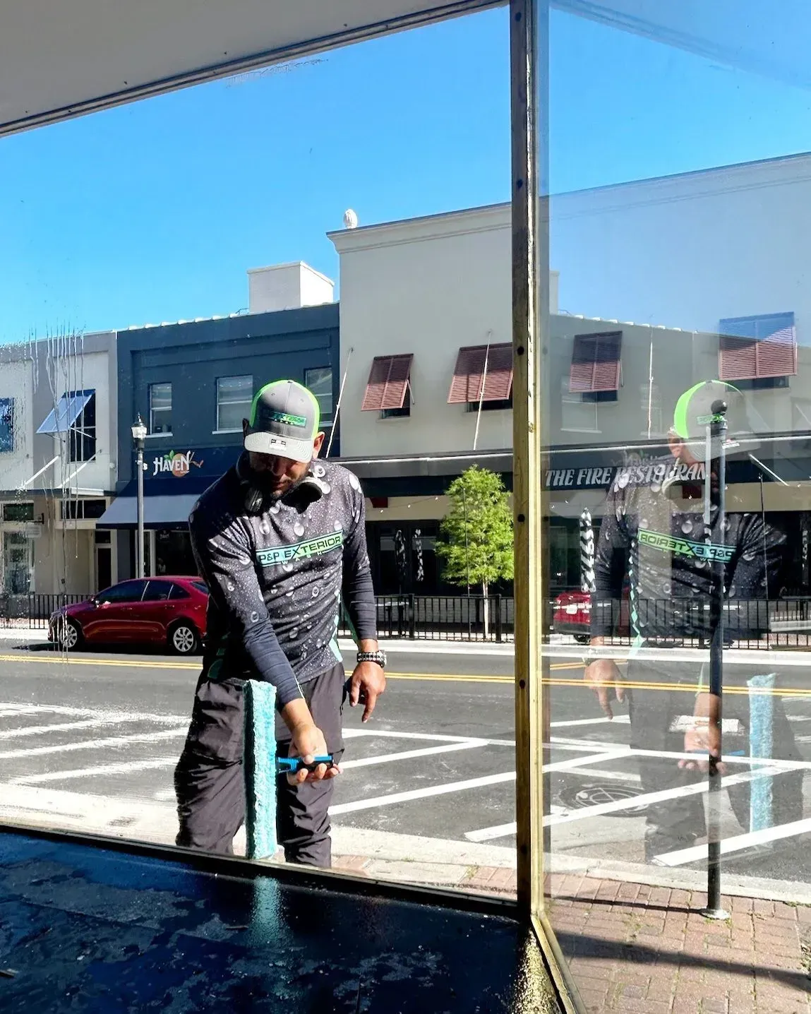 Worker in green hard hat cleaning a glass window outside a storefront.