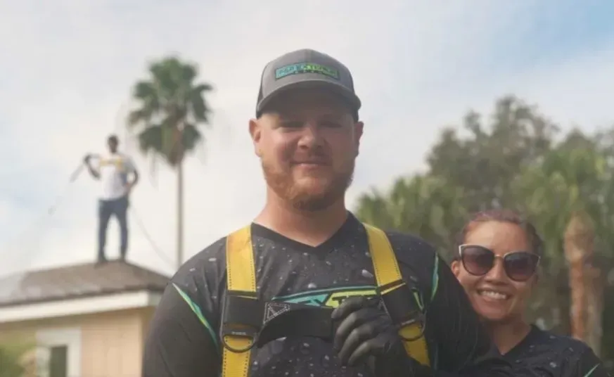 Two smiling people outdoors, one in a black cap and yellow straps, with palm trees in the background
