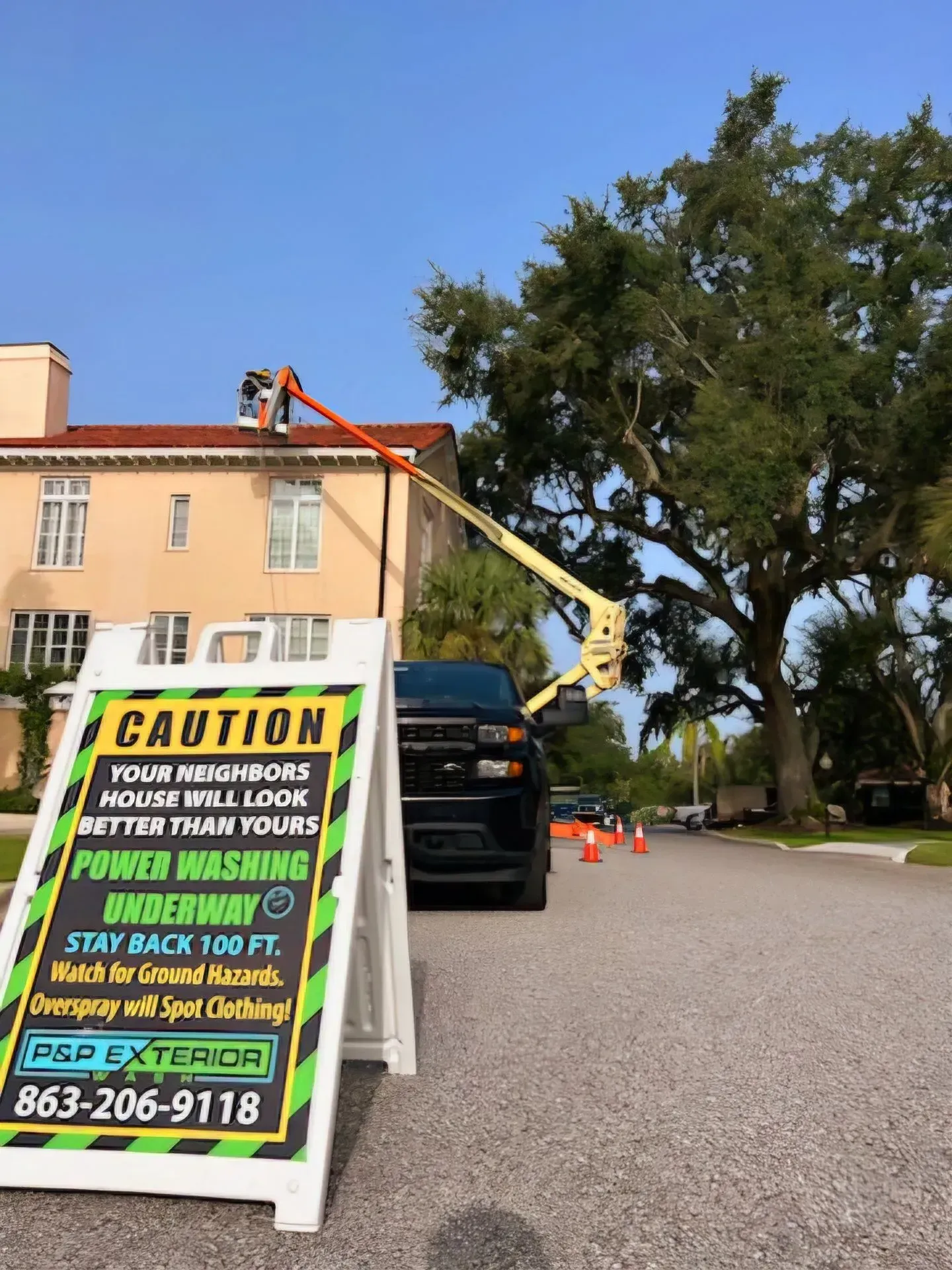 Tree-trimming crew in bucket lifts pruning a large tree beside a peach building and black truck
