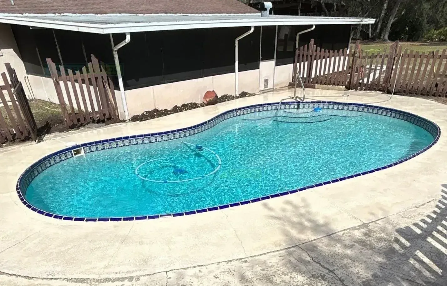 Backyard swimming pool with blue water and a concrete deck, next to a covered patio and wooden fence