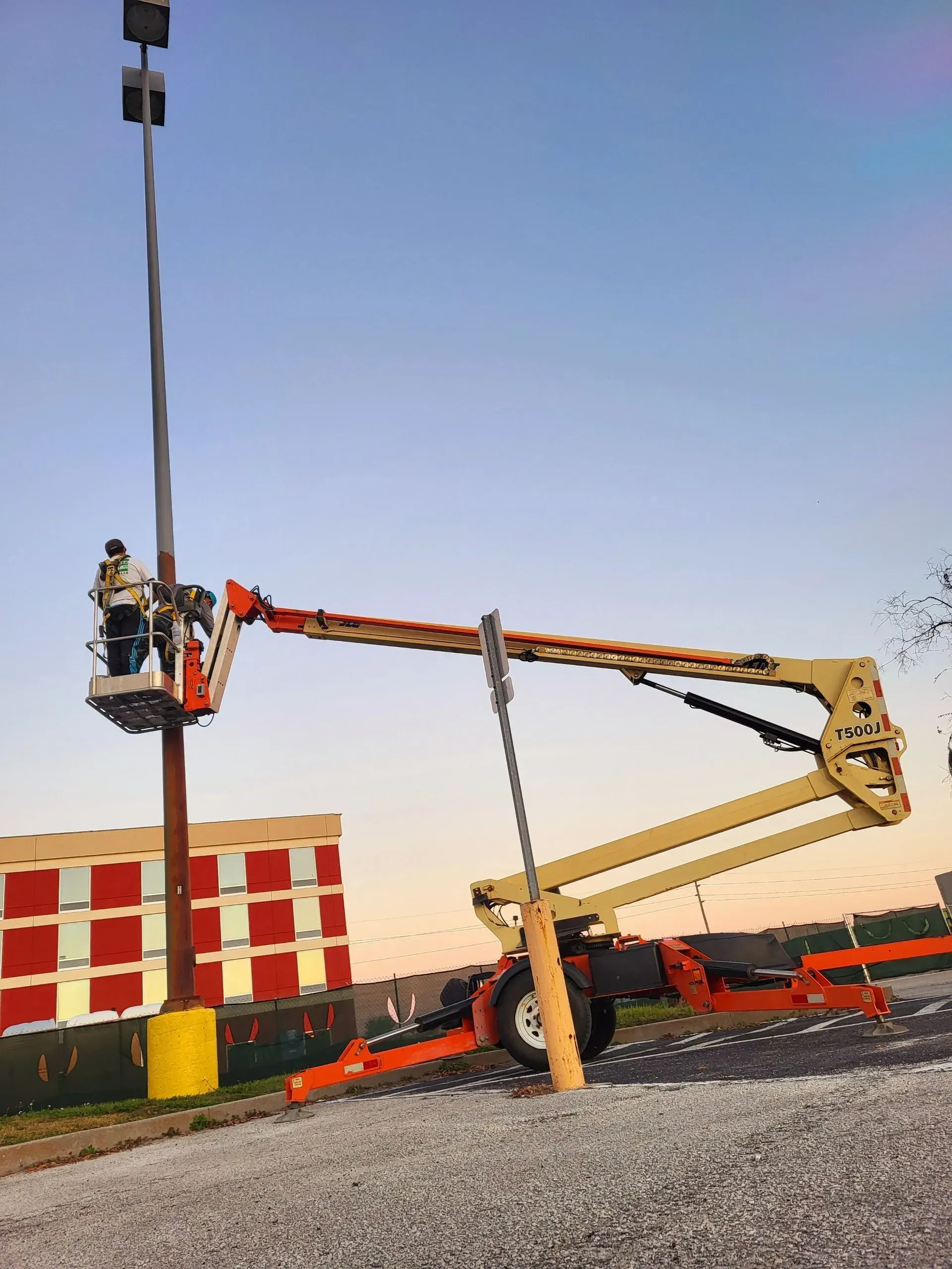Two workers in a cherry picker beside a tall pole at a construction site at sunset