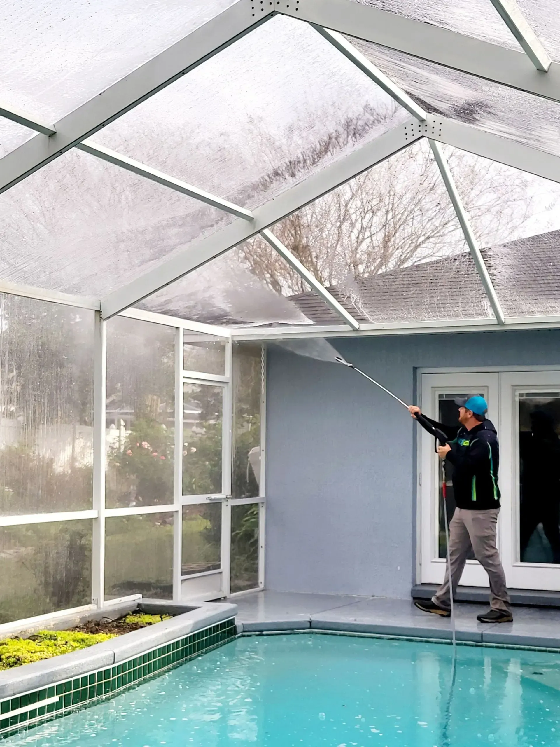 Worker pressure-washing a screened-in pool enclosure beside a turquoise pool