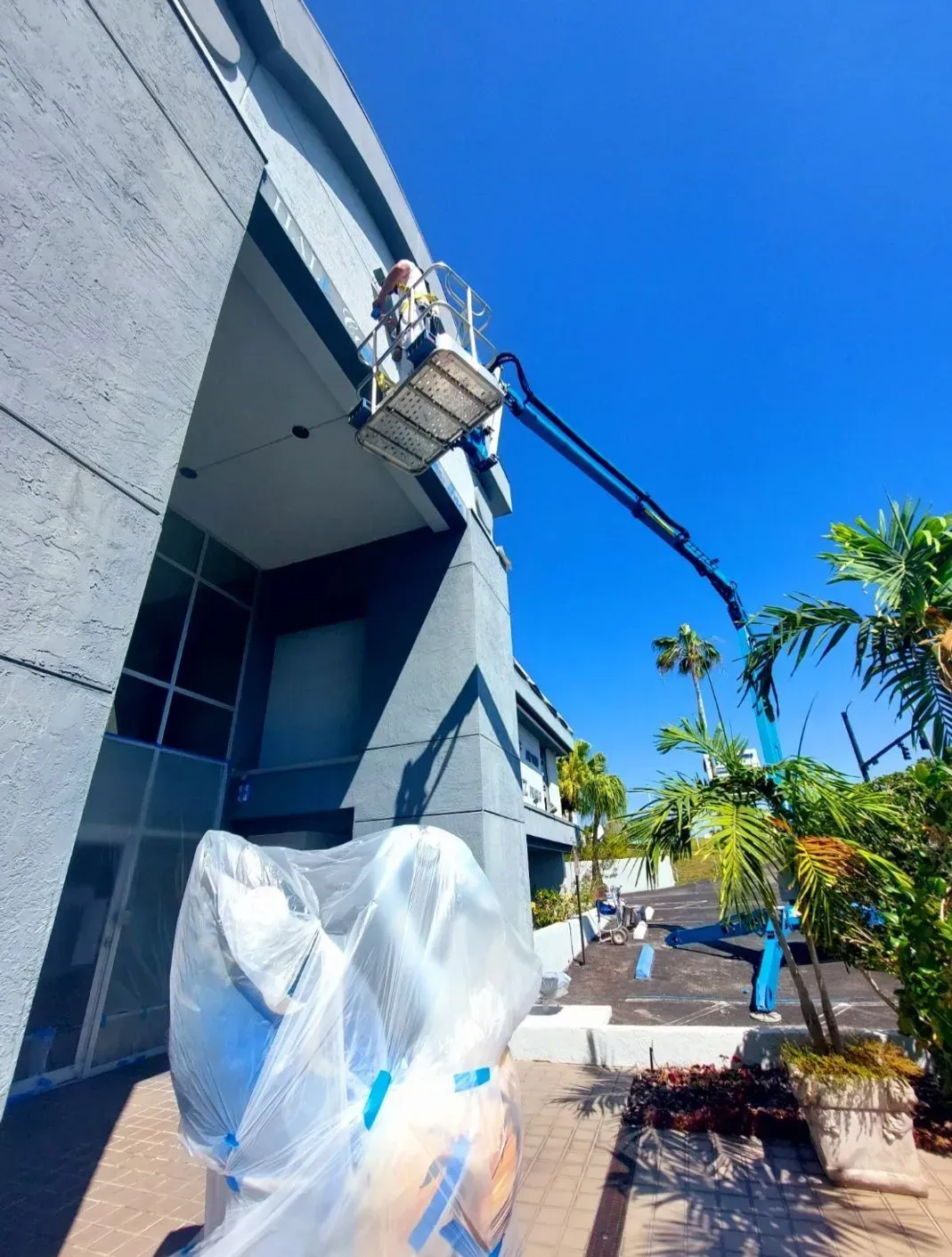 Worker in a lift washing the side of a blue building on a sunny day.